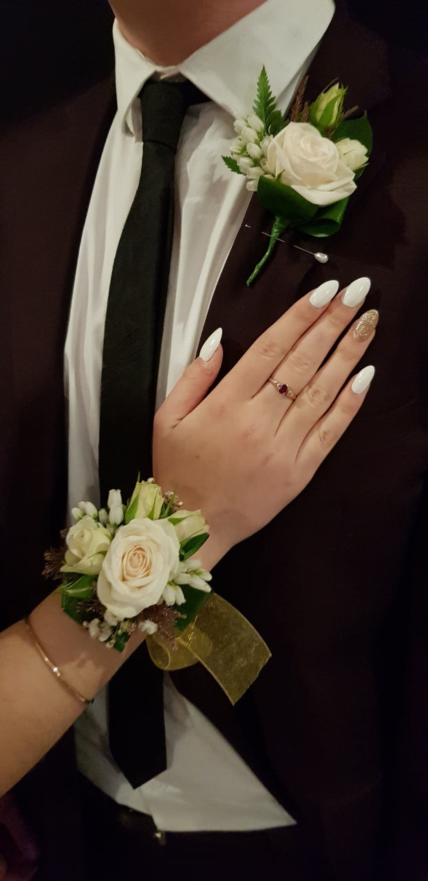A close-up of a wedding or formal event scene showing a man in a dark brown suit, white shirt, and black tie with a white rose boutonniere on his chest, alongside a woman's hand with white manicured nails resting on his chest, wearing a matching white rose wrist corsage.