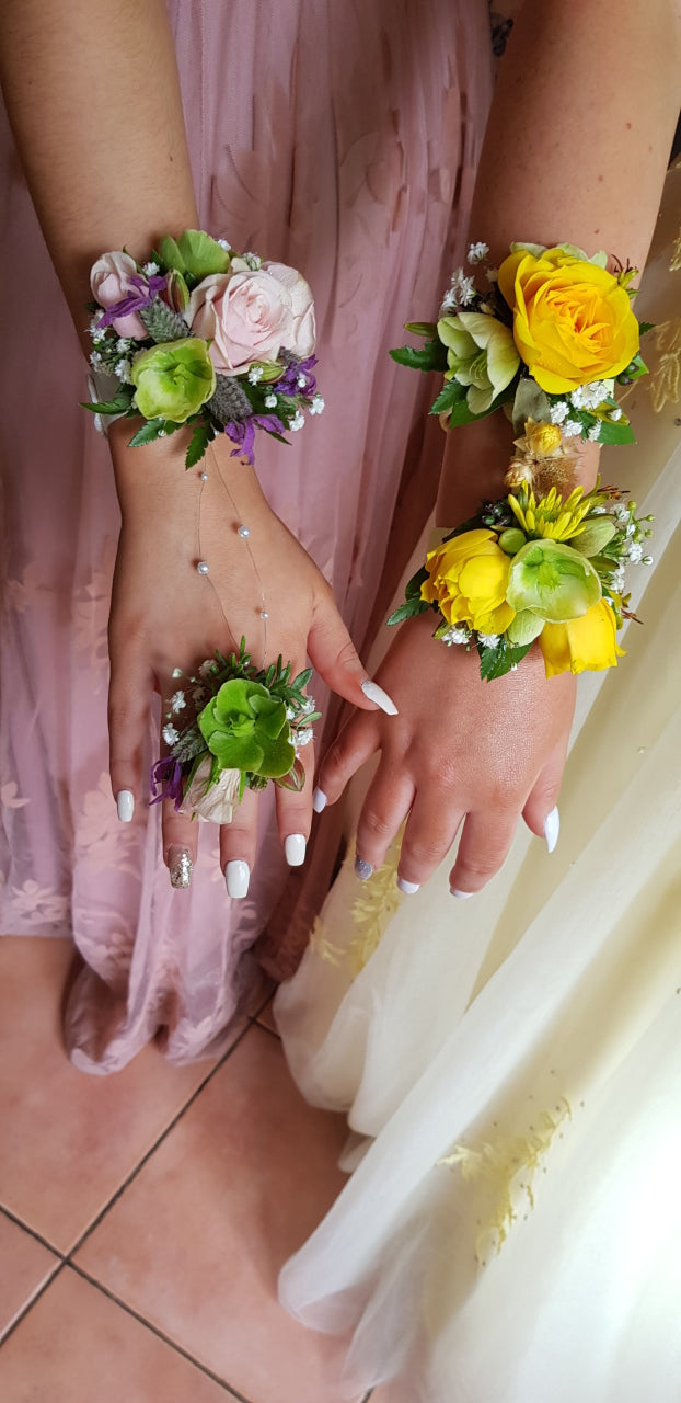 Three wrist corsages and one matching ring worn on hands, styled with a pink dress, white dress, and tiled floor background.