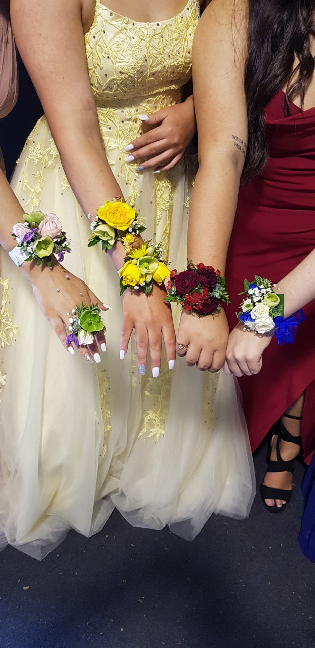 A top-down view of several people sitting together, each wearing a colorful floral wrist corsage. The group includes a person in a red dress, a person in a yellow lace dress, and a person in a white tulle dress, with their hands and arms visible against a dark floor.
