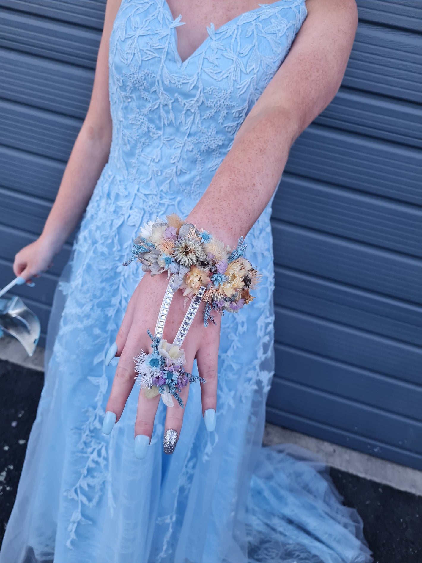 A person wearing a light blue lace dress and matching accessories, including a floral wrist cuff and matching floral ring, standing in front of a blue-gray metal garage door.
