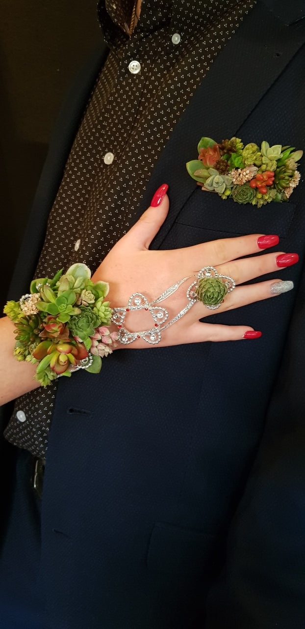 A close-up of a person's hand with red manicured nails, wearing a silver-toned chain bracelet and ring with green succulent floral details, while standing next to someone in a dark navy suit jacket and patterned shirt.