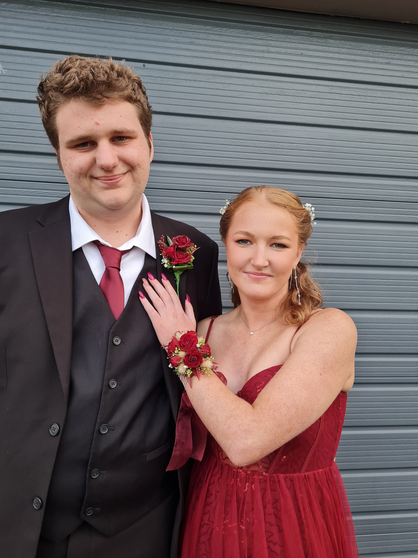 A young man in a black suit and red tie stands next to a young woman in a red strapless dress, both wearing matching red rose wrist and chest florals, in front of a gray metal garage door.