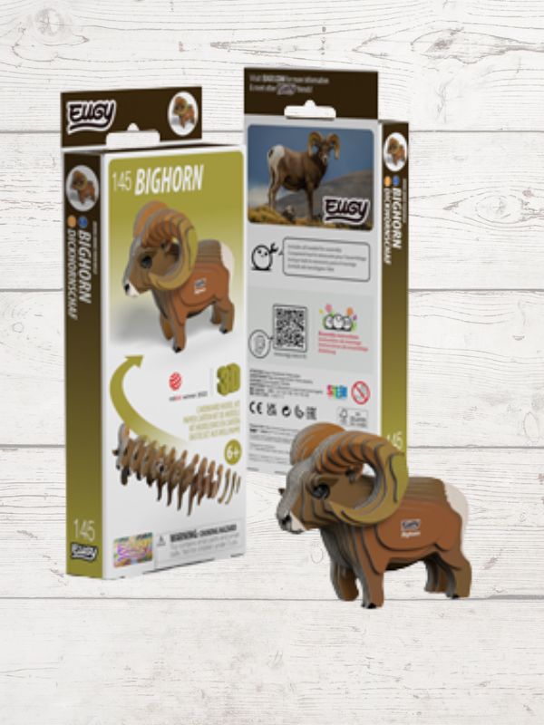 A 1/45 scale wooden bighorn sheep puzzle toy shown next to its retail packaging on a light whitewashed wood background. The product is made of brown wood with a detailed 3D ram figure, and the packaging features green, white, and brown colors with product details and illustrations.