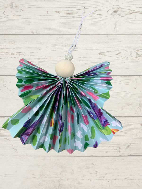 A handmade paper angel ornament with a colorful patterned skirt and a wooden head, hanging against a white wood plank background.