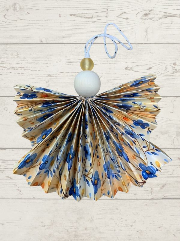 A handmade paper angel Christmas ornament with a pleated fan design, featuring a blue floral pattern on a beige background, hanging from a white wooden bead and light blue string against a white wood plank wall.