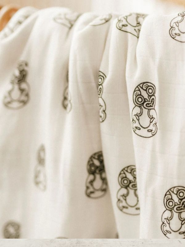A close-up of a white muslin blanket or swaddle with black tribal line-art motifs, draped in soft folds.