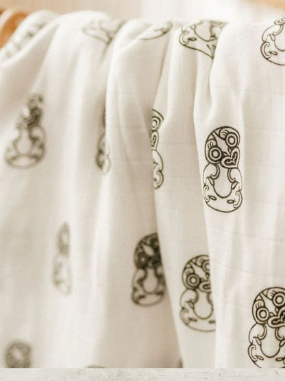 A close-up of a white muslin blanket or swaddle with black tribal line-art motifs, draped in soft folds.