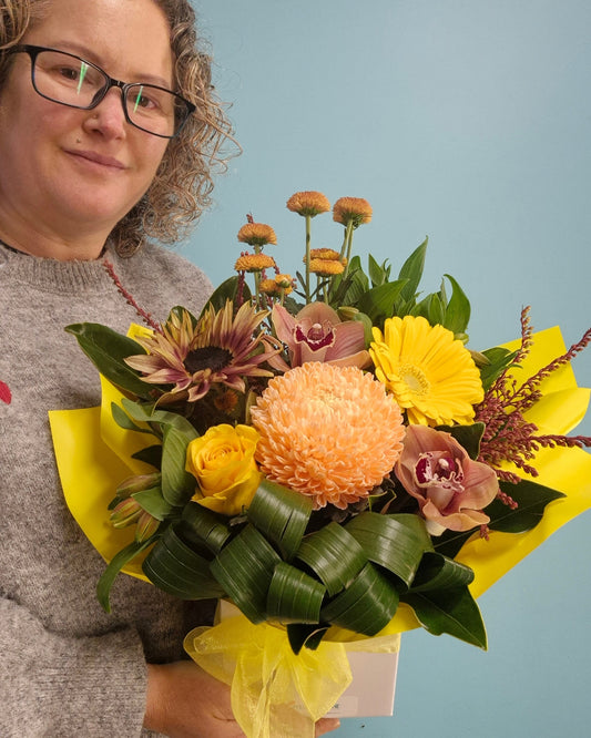 A person wearing glasses and a gray sweater holds a vibrant autumn floral bouquet with yellow gerbera daisies, orange chrysanthemums, purple orchids, and green leaves, wrapped in bright yellow paper against a light blue background.