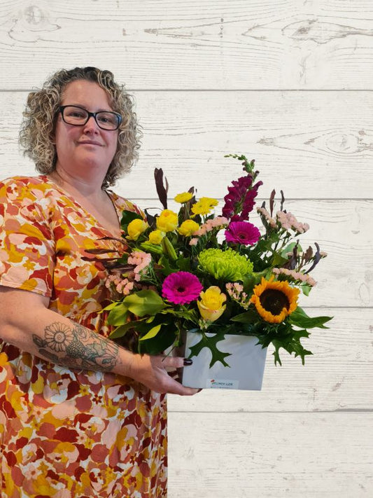 A person holding a floral arrangement with a variety of brightly colored flowers, including pink, yellow, and green, in a container.