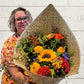 A person holding a bouquet of brightly colored flowers, including yellow, orange, and purple, wrapped in a flax mat with hessian trimmings.