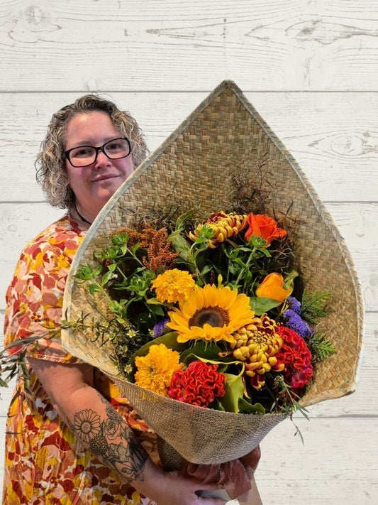 A person holding a bouquet of brightly colored flowers, including yellow, orange, and purple, wrapped in a flax mat with hessian trimmings.
