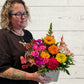 A person holding a floral arrangement with a variety of brightly colored flowers, including pink, yellow, and orange, in a container.