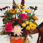 arrangement of bright flowers including yellows, reds and whites in a white box with an orange ribbon