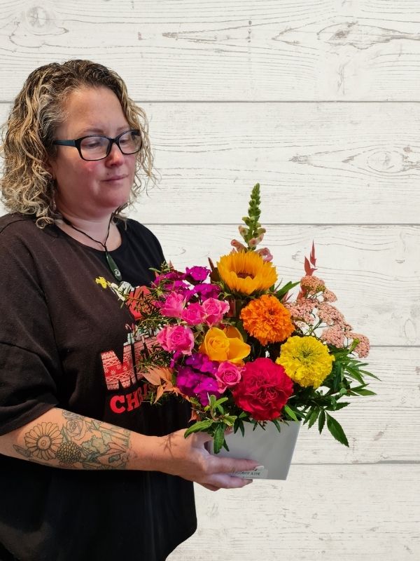 A person holding a bright arrangement of fresh flowers including snapdragons, sunflowers, marigolds, roses made into a white box. 