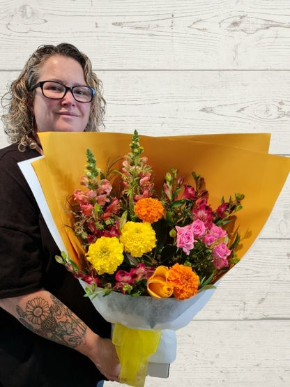 A person holding a bouquet of flowers including snapdragons, marigolds, roses, alstromeria, wrapped in white and yellow with a yellow bow. 
