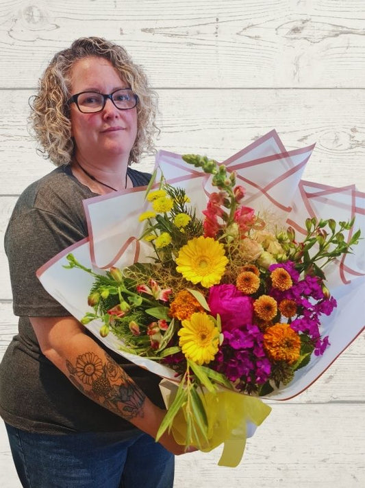A person holding a bouquet of brightly colored flowers, including yellow, orange, and purple, wrapped in mint green paper with a yellow bow.