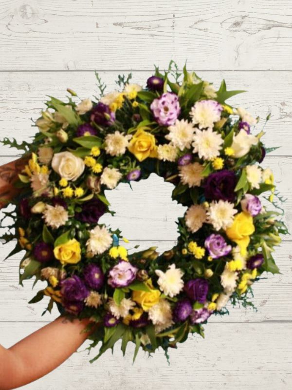 A Bright Sympathy wreath with a mix of purple, yellow, and white flowers, designed to be held in a hand.