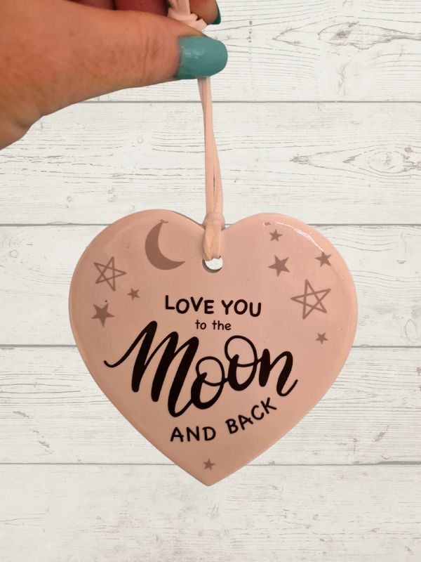 A white ceramic heart-shaped decoration with the phrase 'Love you to the Moon and back' written in black. It is hanging by a string against a wooden background.