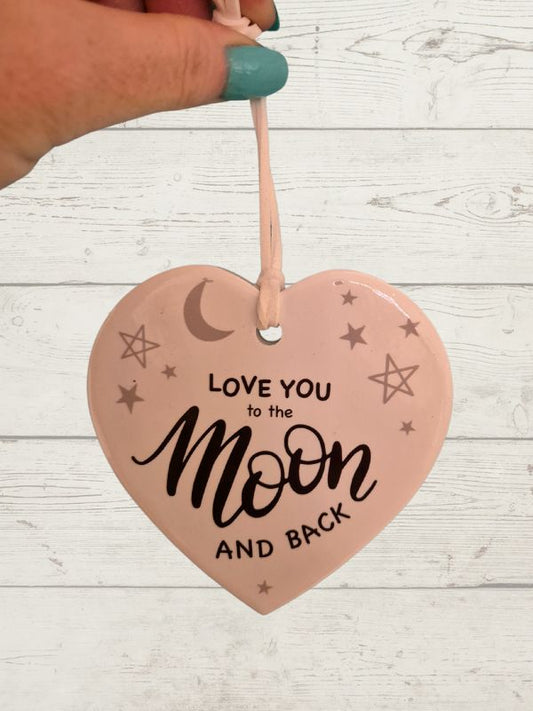 A white ceramic heart-shaped decoration with the phrase 'Love you to the Moon and back' written in black. It is hanging by a string against a wooden background.