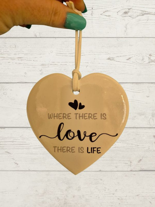 A white ceramic heart-shaped decoration with the text 'WHERE THERE IS LOVE THERE IS LIFE' printed on it, hanging against a wooden background.