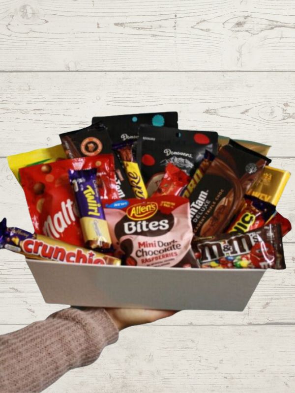 A gift box filled with various chocolate treats and sweets, held in a person's hand against a wooden background.