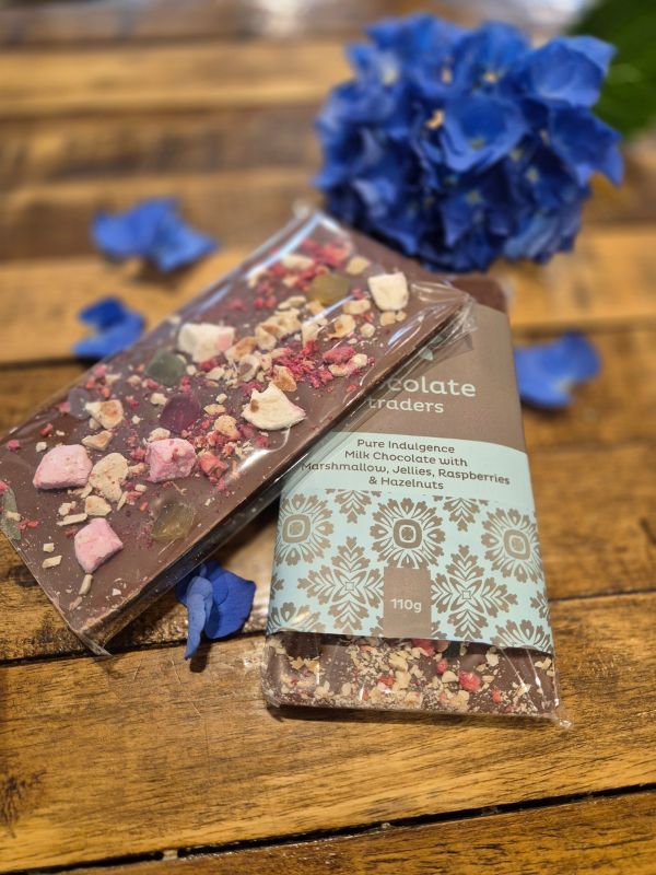 A chocolate bar with decorative toppings, placed on a wooden surface next to a flower, with the brand 'Chocolate Traders' visible on the packaging.