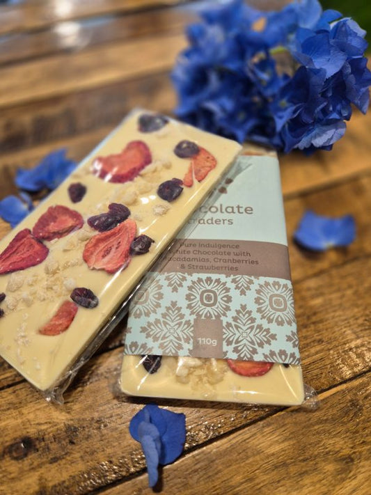 A chocolate bar with decorative toppings, placed on a wooden surface next to a flower, with the brand 'Chocolate Traders' visible on the packaging.