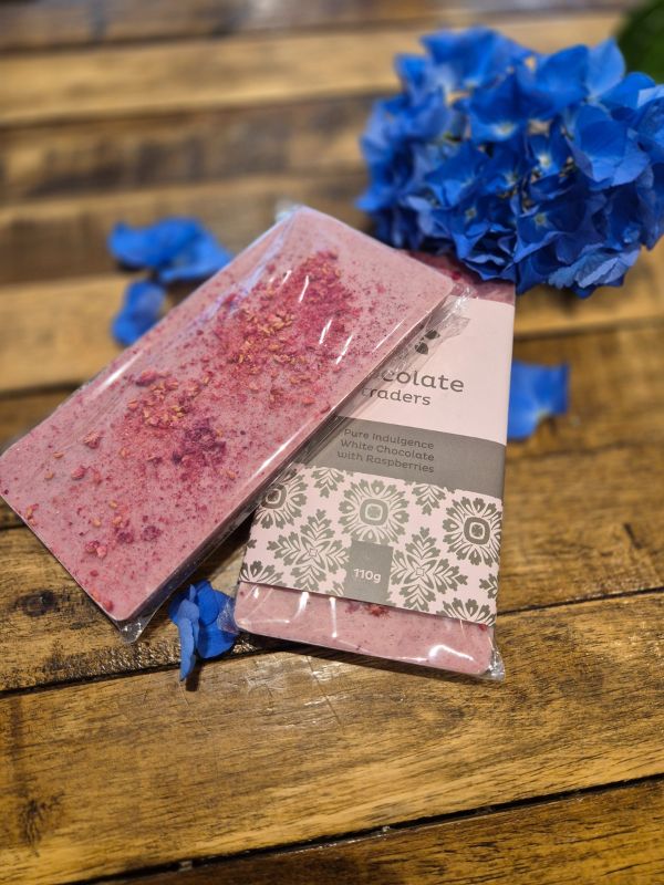 A chocolate bar with decorative toppings, placed on a wooden surface next to a flower, with the brand 'Chocolate Traders' visible on the packaging.