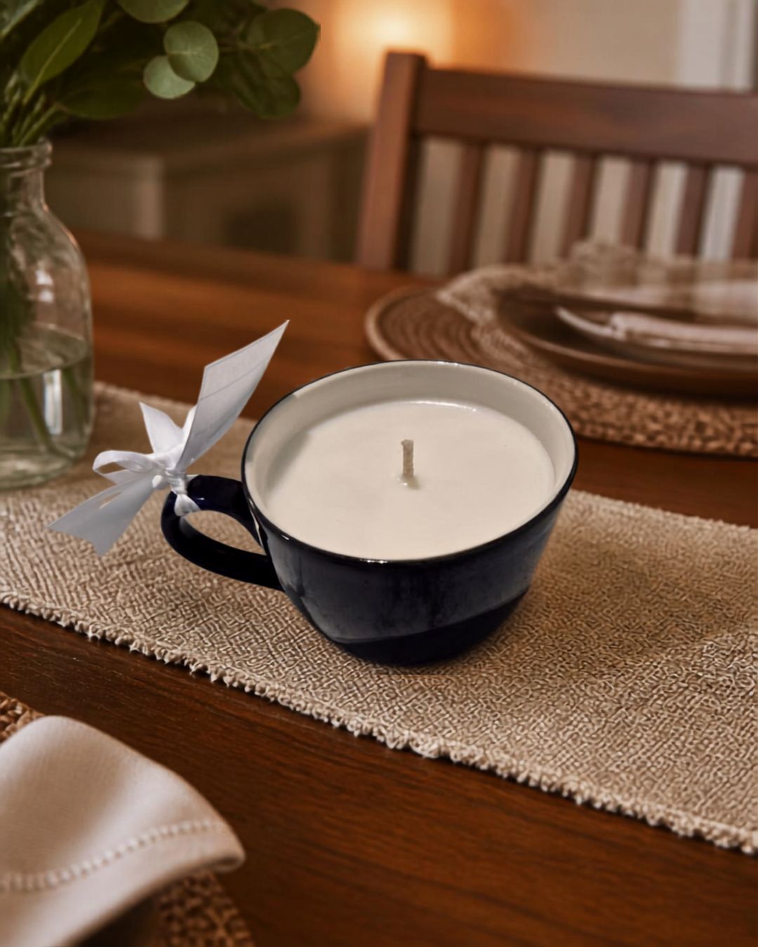 A navy blue ceramic mug candle with white wax sits on a woven beige placemat on a wooden dining table. A white paper airplane is tied to the handle, and a blurred wooden chair, plates, and a glass vase with greenery are in the background.