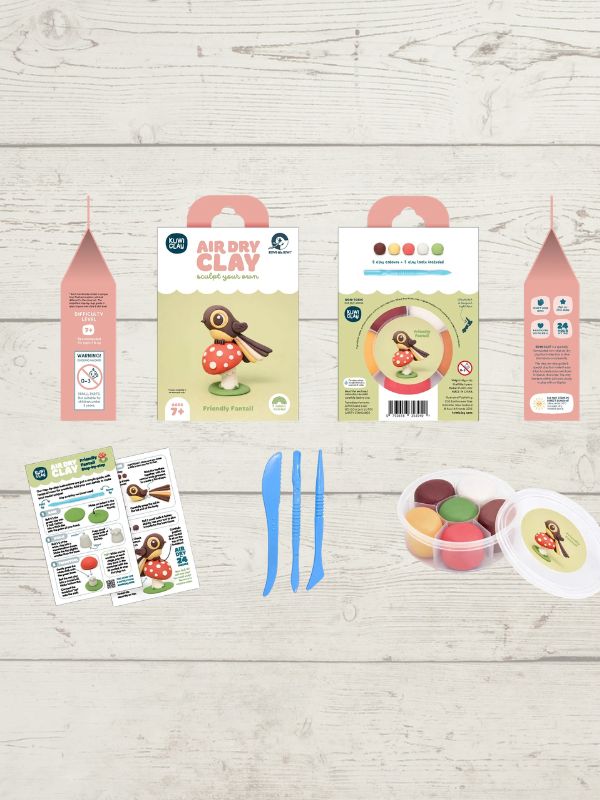 Air Dry Clay craft kit components laid out on a light wood background, including three pink and green cardboard packaging boxes, two instruction sheets, two blue plastic knives, and a clear plastic tub of colorful clay with a white lid.