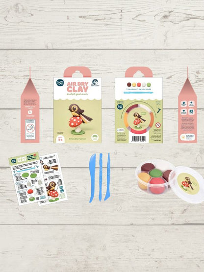 Air Dry Clay craft kit components laid out on a light wood background, including three pink and green cardboard packaging boxes, two instruction sheets, two blue plastic knives, and a clear plastic tub of colorful clay with a white lid.