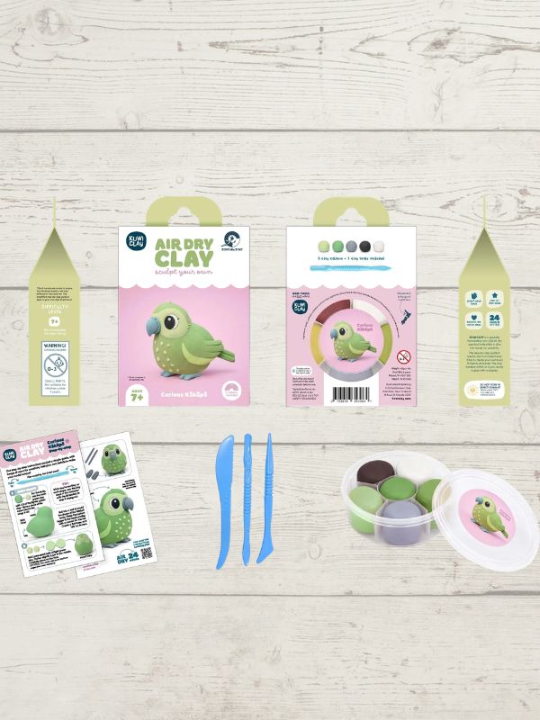 A bird-themed air dry clay kit laid out on a light wood background, including two main product boxes with green parrot illustrations, three small clay pots, blue sculpting tools, instruction sheets, and a lid with a parrot logo.