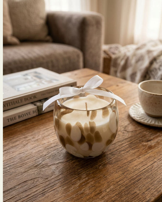 A glass jar candle with a white wax body, brown leaf-like swirls, and a white ribbon bow, sitting on a wooden table in a cozy living room setting.