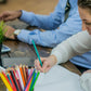 An adult is coloring a page in a coloring book using a pencil, with a cup of coloring pencils and a sharpener beside it.