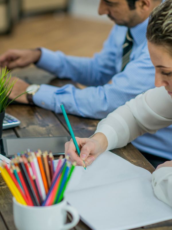 An adult is coloring a page in a coloring book using a pencil, with a cup of coloring pencils and a sharpener beside it.