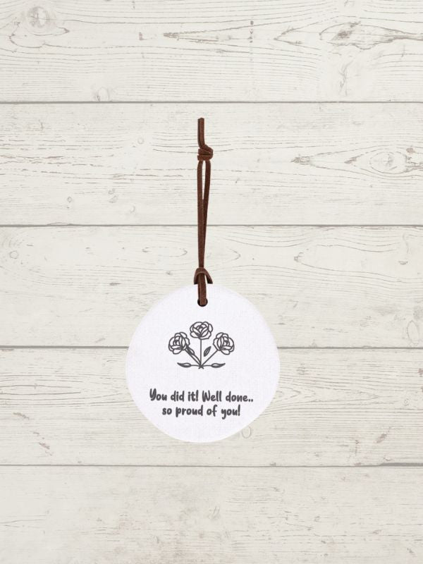A white circular hanging ornament with a brown hanging cord, featuring a black line drawing of three roses and the printed message 'You did it! Well done.