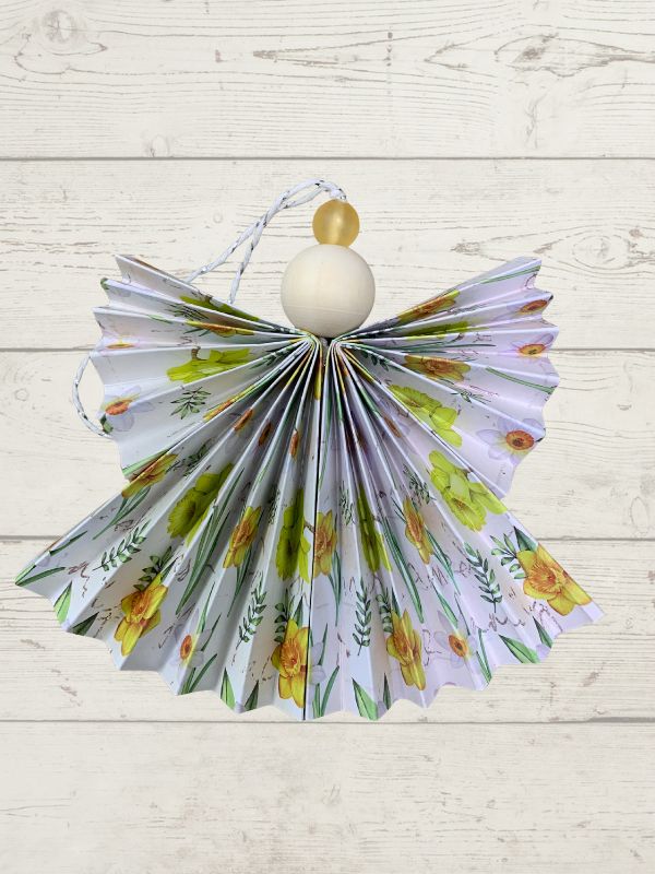 A decorative hanging ornament shaped like a paper angel with a pleated white wings patterned with yellow flowers and green leaves, featuring a white wooden body and gold bead accents, set against a light wood plank background.