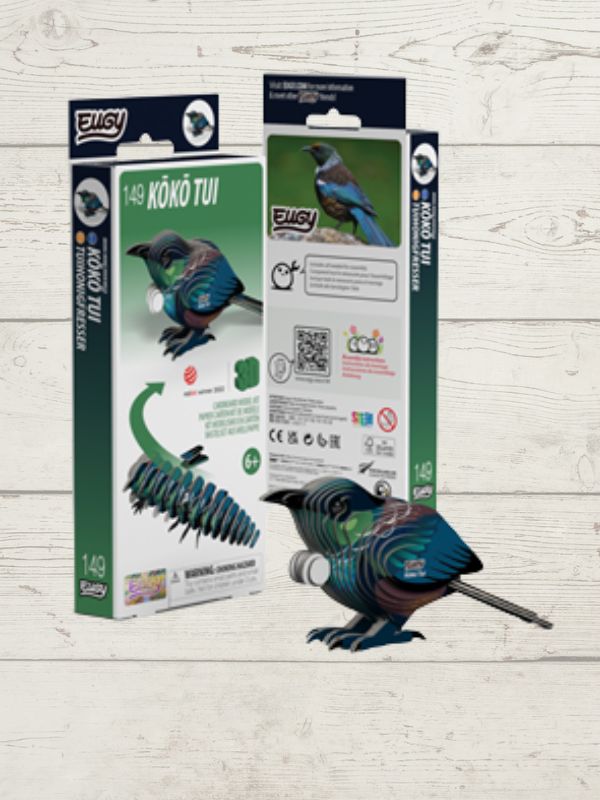 A 14-piece wooden Kōkō Tui puzzle set shown with its retail packaging on a light whitewashed wood background. The assembled bird is in front of the boxes, and the packaging features green and white panels with illustrations of the bird and the wooden pieces.