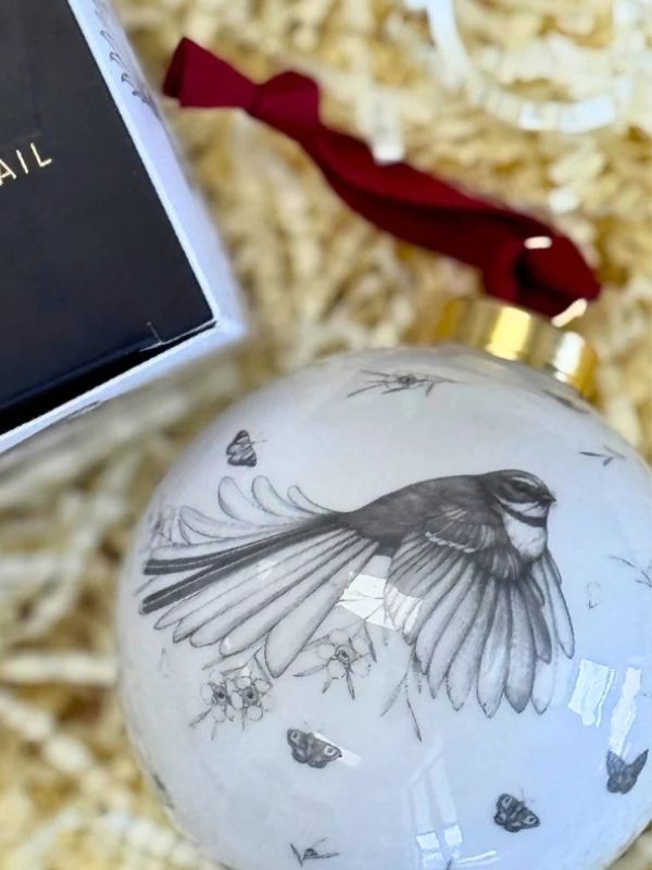 A round white Christmas ornament featuring a detailed black and white sketch of a bird in flight, decorated with small butterfly motifs. The ornament has a gold cap and a red ribbon, shown next to its black gift box on a bed of beige paper shred.