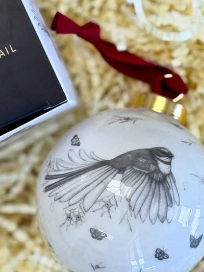 A round white Christmas ornament featuring a detailed black and white sketch of a bird in flight, decorated with small butterfly motifs. The ornament has a gold cap and a red ribbon, shown next to its black gift box on a bed of beige paper shred.