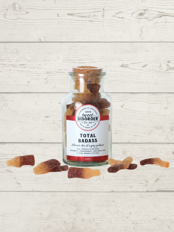 A clear glass jar filled with brown gummy candies, labeled 'Sweet Disorder' and 'Total Badass,' placed on a white wood plank background with a few gummies scattered around the base.