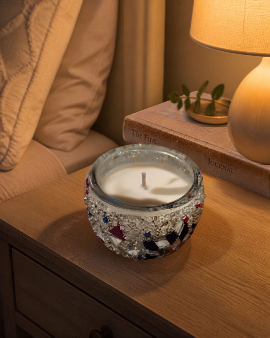 A lit white candle in a clear glass holder decorated with a silver and multicolor mosaic tile pattern, styled on a wooden bedside table next to a lamp and book.