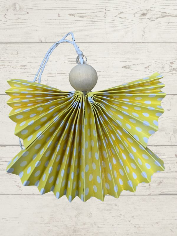 A handmade angel Christmas ornament featuring a yellow and light blue polka-dotted paper fan body, a light wood ball torso, and a light blue hanging string, set against a light wood plank background.
