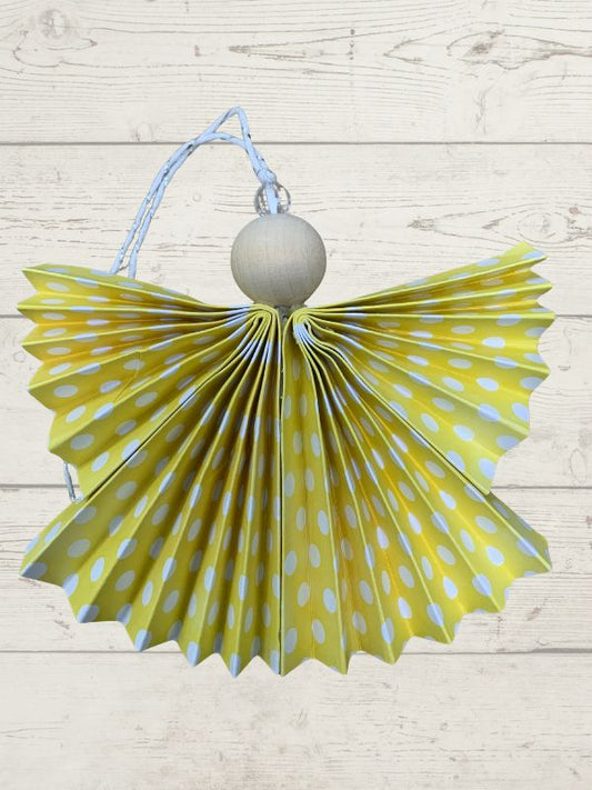 A handmade angel Christmas ornament featuring a yellow and light blue polka-dotted paper fan body, a light wood ball torso, and a light blue hanging string, set against a light wood plank background.