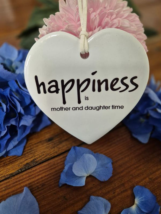 A ceramic heart-shaped plaque with the word 'Happiness' written on it, displayed on a wooden surface surrounded by blue flower petals and a pink flower.
