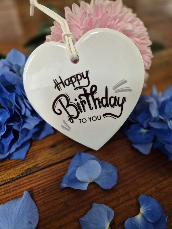 A white ceramic heart-shaped plaque with the words 'Happy Birthday' and a small pink flower attached to it, placed on a wooden surface surrounded by blue ceramic flowers.