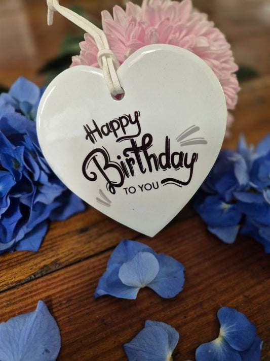 A white ceramic heart-shaped plaque with the words 'Happy Birthday' and a small pink flower attached to it, placed on a wooden surface surrounded by blue ceramic flowers.