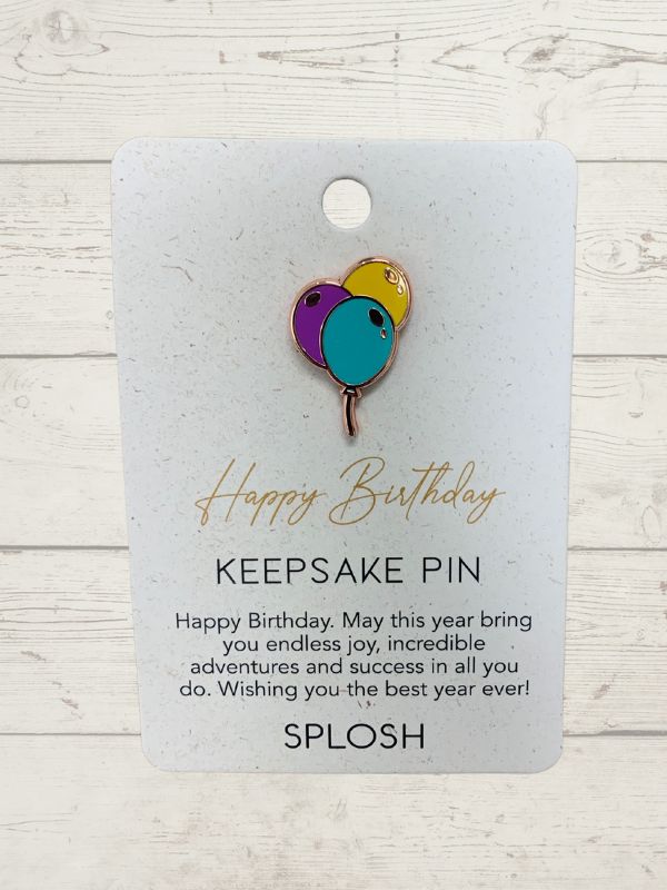A keepsake pin with a multicolored design, featuring a balloon and a heart, attached to a card that reads 'Happy Birthday Keepsake Pin'.