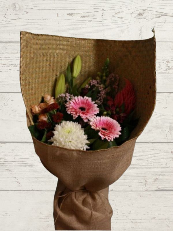 A bouquet of flowers including pink, white, and green flowers, wrapped in a brown paper with hessian trimmings, set against a wooden background.