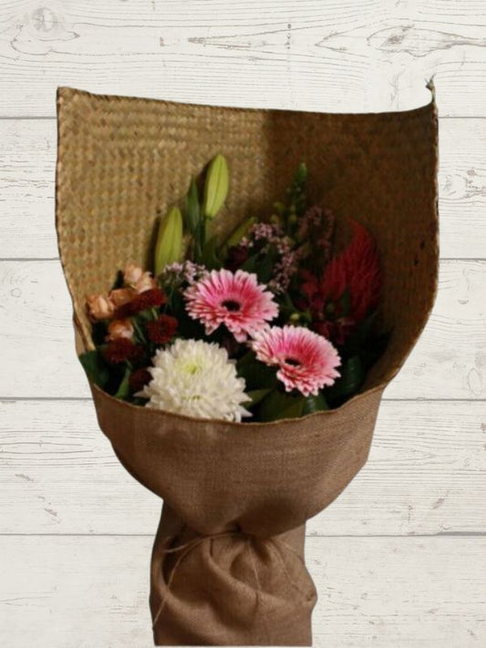 A bouquet of flowers including pink, white, and green flowers, wrapped in a brown paper with hessian trimmings, set against a wooden background.